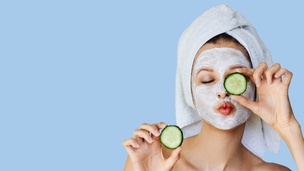 Beautiful young woman with facial mask on her face holding slices of cucumber. Skin care and treatment, spa, natural beauty and cosmetology concept.